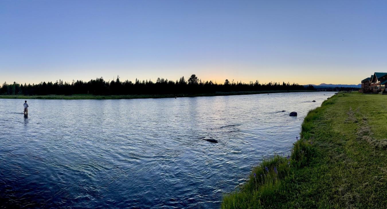Fly fisherman wading the Henry's Fork of the Snake River at dusk with Anglers Lodge visible on the bank in Island Park Idaho